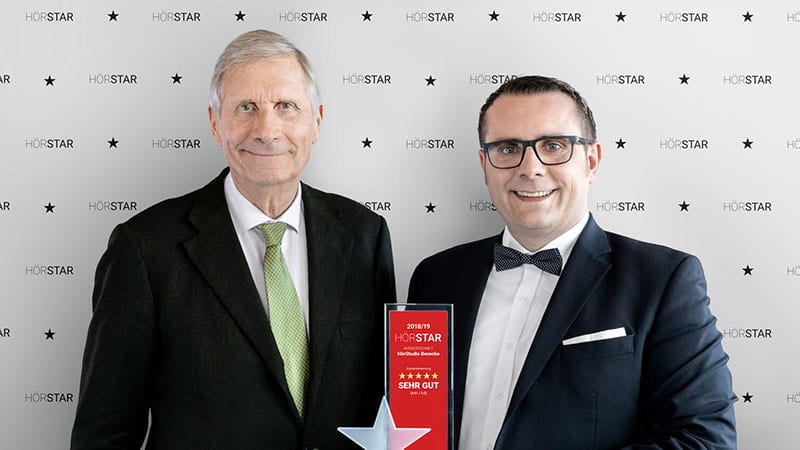 Zwei Maenner laecheln im Anzug vor HORSTAR Wand mit roter Trophäe
